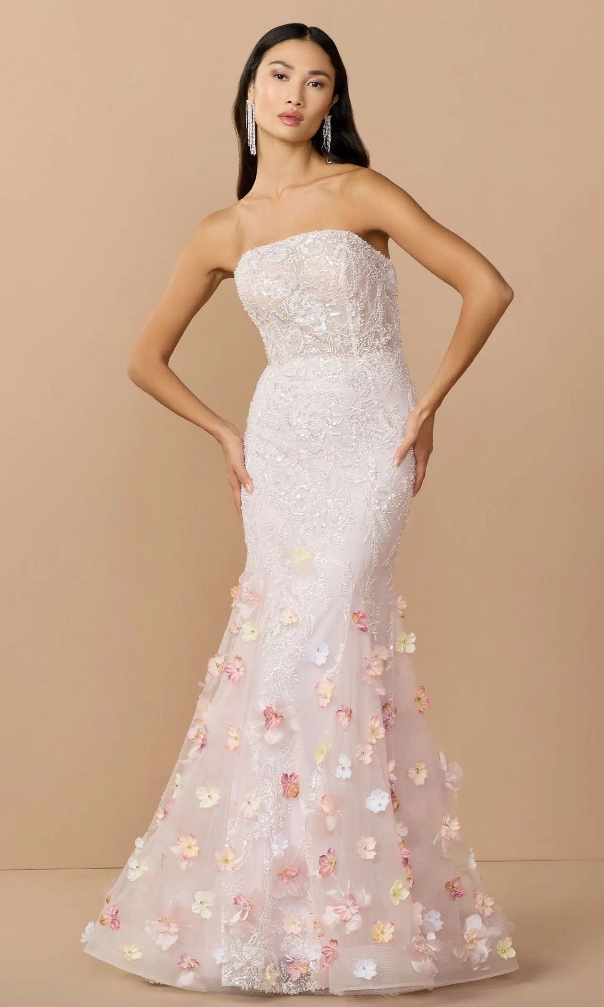 A woman poses in the Mac Duggal 21147 long formal dress, a strapless mermaid gown with intricate lace and pastel floral appliqués on the skirt, set against a plain beige background.